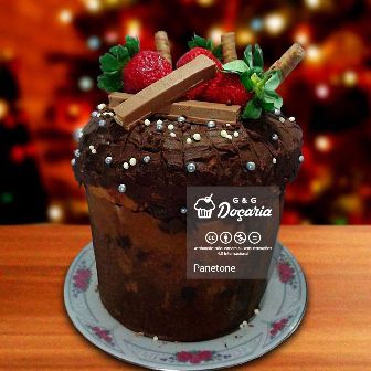 Luzes de Natal e uma mesa com um panetone recheado de brigadeiro gourmet enfeitado com confeitos crocantes prateados, barrinhas de kit kat, morangos e biscoitos